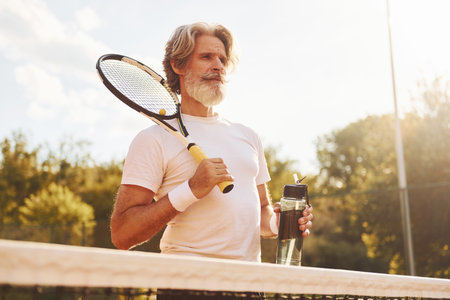 Holding racket. Senior modern stylish man with racket outdoors on tennis court at daytimeの写真素材