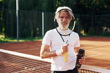 Holding bottle of water. Senior modern stylish man with racket outdoors on tennis court at daytimeの写真素材