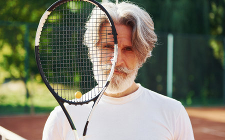 Holding racket. Senior modern stylish man with racket outdoors on tennis court at daytimeの写真素材