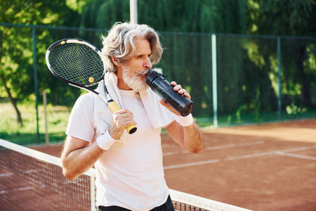 Holding bottle of water. Senior modern stylish man with racket outdoors on tennis court at daytimeの写真素材