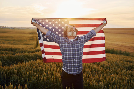 Holding USA flag in hands. Patriotic senior stylish man with grey hair and beard on the agricultural...