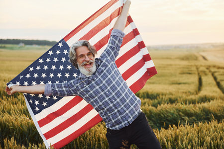 Holding USA flag in hands. Patriotic senior stylish man with grey hair and beard on the agricultural fieldの写真素材