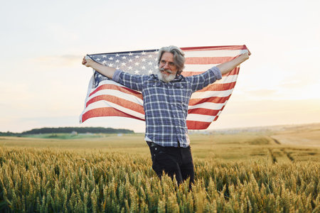 Holding USA flag in hands. Patriotic senior stylish man with grey hair and beard on the agricultural fieldの写真素材