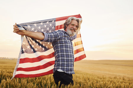 Holding USA flag in hands. Patriotic senior stylish man with grey hair and beard on the agricultural fieldの写真素材