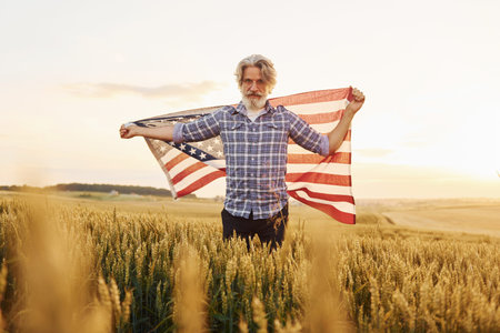 Holding USA flag in hands. Patriotic senior stylish man with grey hair and beard on the agricultural...