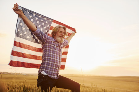 Holding USA flag in hands. Patriotic senior stylish man with grey hair and beard on the agricultural fieldの写真素材
