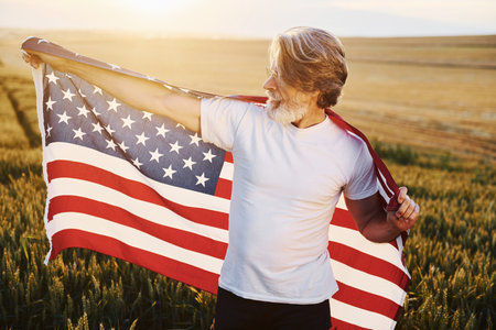 Conception of freedom. Holding USA flag. Senior stylish man with grey hair and beard on the agricultural field with harvestの写真素材