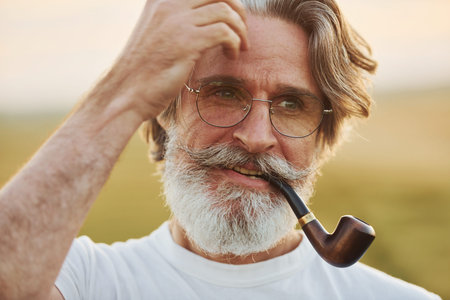 Portrait of senior stylish man with grey hair and beard that standing outdoors on field at sunny day...