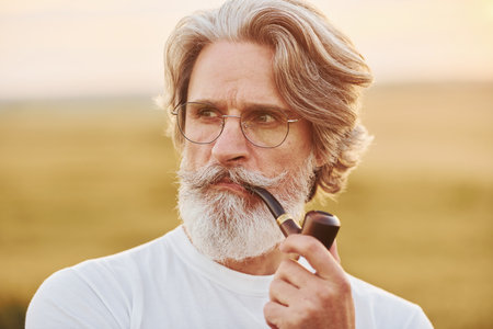 Portrait of senior stylish man with grey hair and beard that standing outdoors on field at sunny day and smokingの写真素材