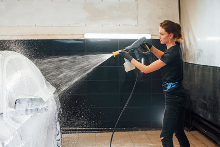 Using high pressure water. Modern black automobile get cleaned by woman inside of car wash stationの写真素材