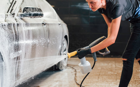 Using high pressure water. Modern black automobile get cleaned by woman inside of car wash stationの写真素材
