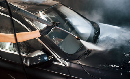 Using equipment with high pressure water. Modern black automobile get cleaned by woman inside of car wash stationの写真素材