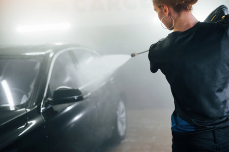 Using equipment with high pressure water. Modern black automobile get cleaned by woman inside of car wash stationの写真素材
