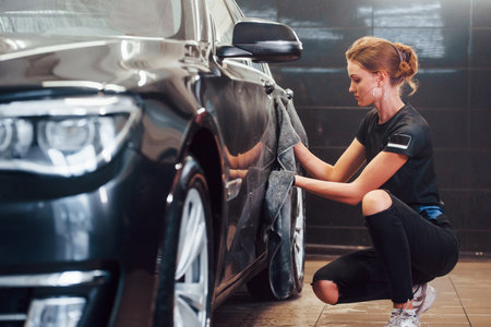 Wipes surface by using towel. Modern black automobile get cleaned by woman inside of car wash stationの写真素材
