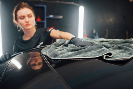 Wipes surface by using towel. Modern black automobile get cleaned by woman inside of car wash stationの写真素材