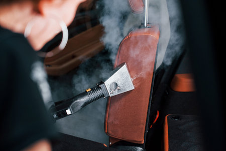 Uses steam cleaner. Modern black automobile is in service by woman inside of car wash station