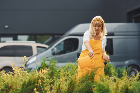 Senior woman in yellow colored uniform is in the garden at daytime watering plantsの写真素材