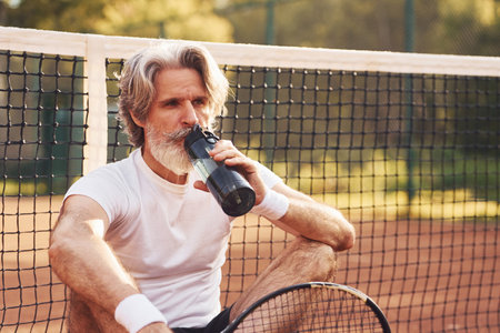 Taking break and drinking water. Senior stylish man in white shirt and black sportive shorts on tennis courtの写真素材