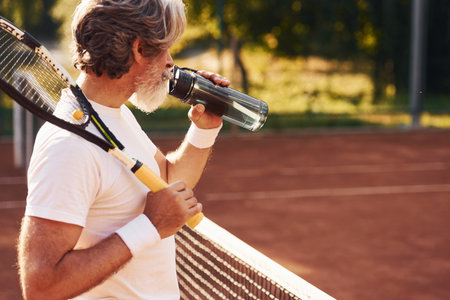 Taking break and drinking water. Senior stylish man in white shirt and black sportive shorts on tennis courtの写真素材