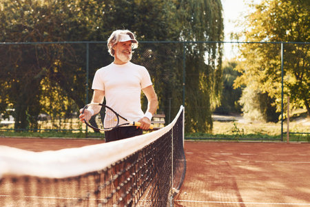 Beautiful sunlight. Senior stylish man in white shirt and black sportive shorts on tennis courtの写真素材