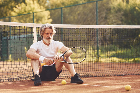 Sitting and taking a break. Senior stylish man in white shirt and black sportive shorts on tennis courtの写真素材