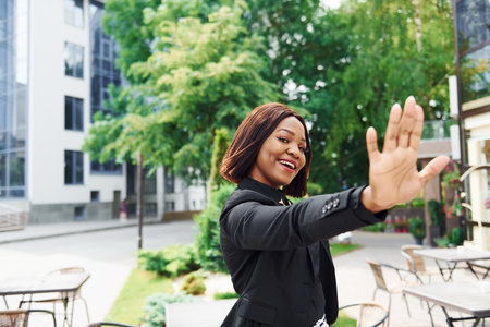 High five gesture. Young afro american woman in fashionable clothes outdoors in the city near green trees and against business buildingの写真素材