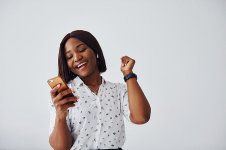 Positive african american woman in white shirt standing indoors against white wall with phone in handの写真素材