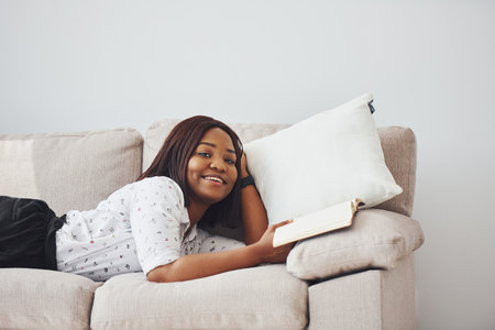 Positive african american woman in white shirt lying down on sofa with notepad in handsの写真素材