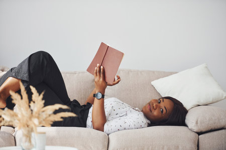 Positive african american woman in white shirt lying down on sofa with notepad in handsの写真素材