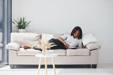 Positive african american woman in white shirt lying down on sofa with notepad in handsの写真素材