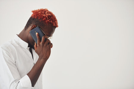 Young african american man in white formal clothes talking by the phone against wall indoorsの写真素材