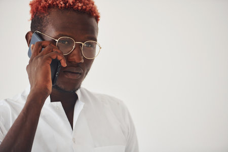 Young african american man in white formal clothes talking by the phone against wall indoorsの写真素材