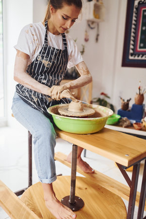 Young female ceramist working by using pottery wheel indoors and making handmade clay productの写真素材