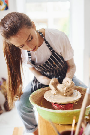 Young female ceramist working by using pottery wheel indoors and making handmade clay productの写真素材