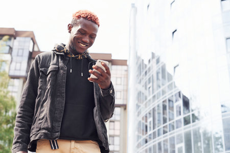 Holding phone. Young african american man in black jacket outdoors in the city standing against modern business buildingの写真素材