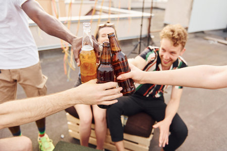 Doing cheers by bottles with beer. Group of young people in casual clothes have a party at rooftop together at daytimeの写真素材