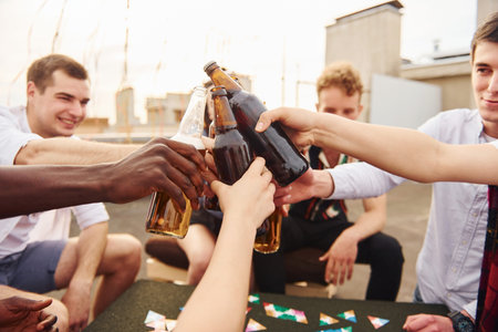Doing cheers by bottles with beer. Group of young people in casual clothes have a party at rooftop...