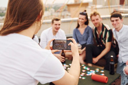 Girl doing photo when people playing card game. Group of young people in casual clothes have a party at rooftop together at daytimeの写真素材