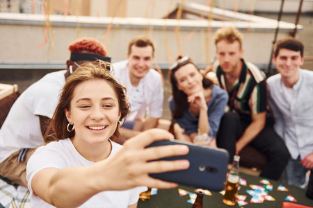 Girl doing photo when people playing card game. Group of young people in casual clothes have a party at rooftop together at daytimeの写真素材