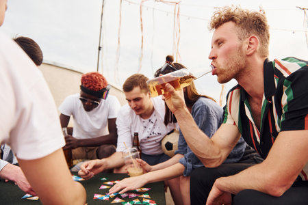 Playing card game. Group of young people in casual clothes have a party at rooftop together at daytimeの写真素材
