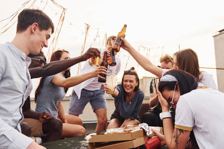 Doing cheers by bottles with beer. Group of young people in casual clothes have a party at rooftop together at daytimeの写真素材