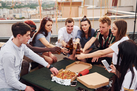 Doing cheers by bottles with beer. Group of young people in casual clothes have a party at rooftop together at daytimeの写真素材
