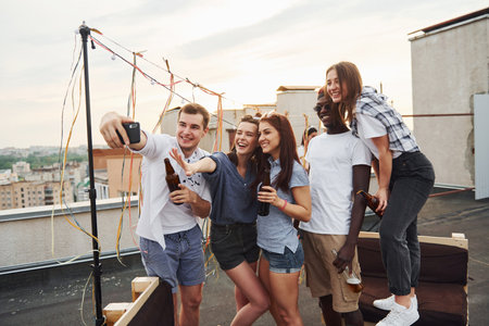 Group of young people in casual clothes have a party at rooftop together at daytime and doing selfie by phoneの写真素材