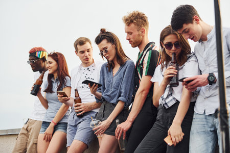 Standing with phones and alcohol in hands. Group of young people in casual clothes have a party at rooftop together at daytimeの写真素材