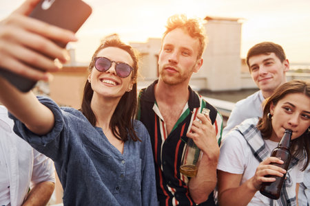 Taking selfie. Beautiful sunshine. Group of young people in casual clothes have a party at rooftop together at daytimeの写真素材