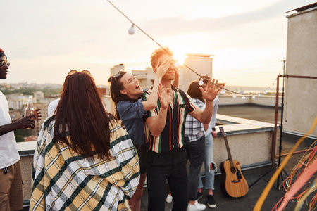 Playing game. Mans eyes covered by hands. Group of young people in casual clothes have a party at rooftop together at daytimeの写真素材