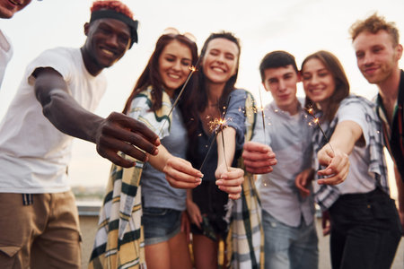 With sparklers in hands. Group of young people in casual clothes have a party at rooftop together at...