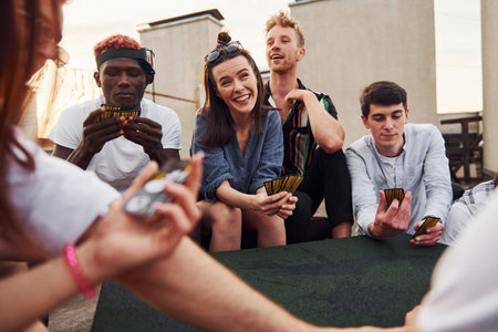 Sitting by table and playing card game. Group of young people in casual clothes have a party at rooftop together at daytimeの写真素材