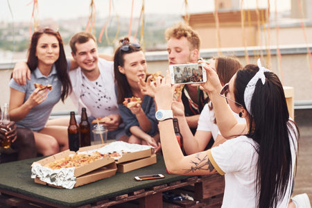 Making photo. Delicious pizza. Group of young people in casual clothes have a party at rooftop together at daytimeの写真素材