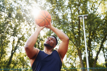 Beautiful green trees on background. African american man plays basketball on the court outdoorsの写真素材